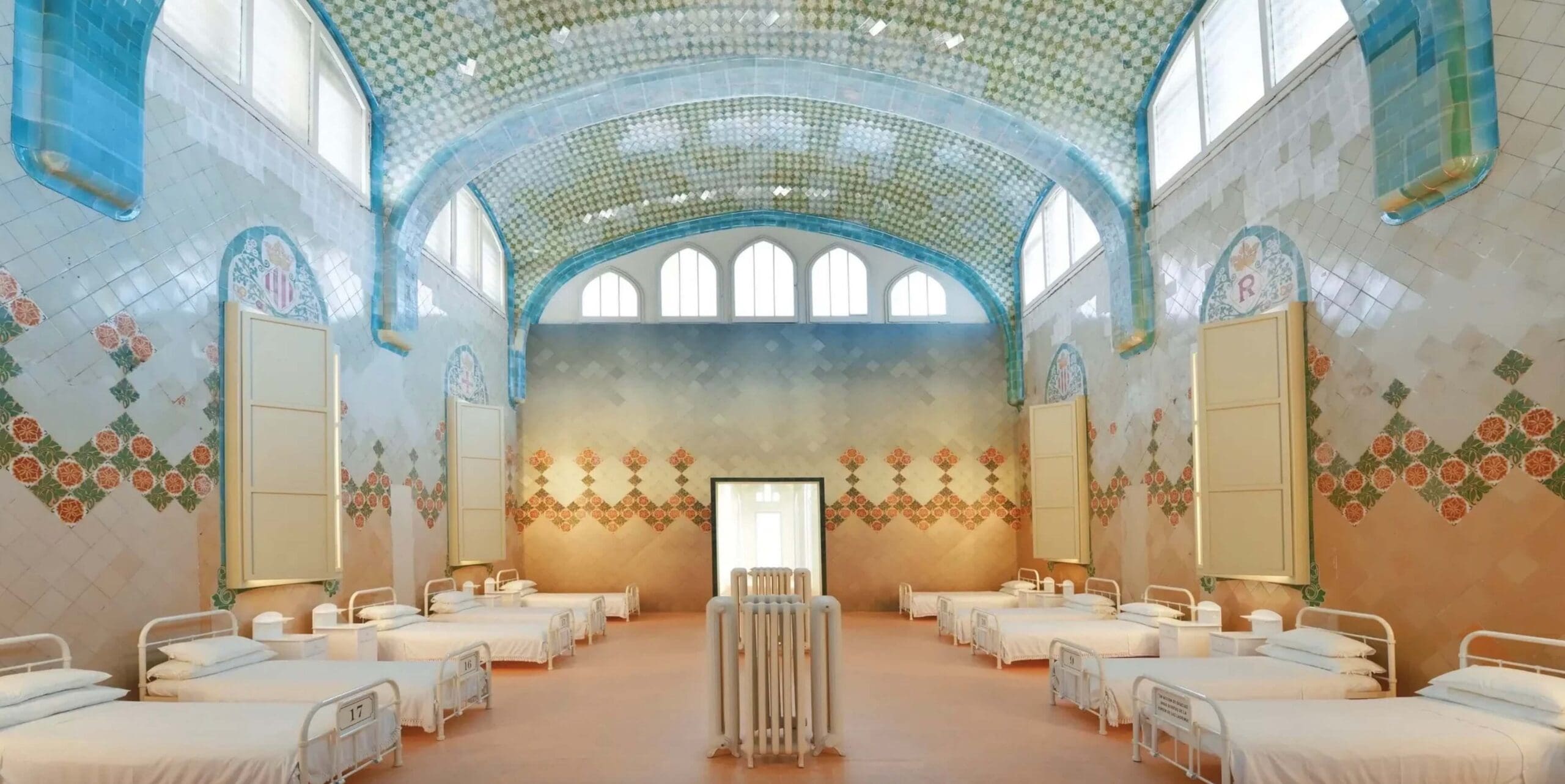 A patient ward at Hospital de Sant Pau in Barcelona natural light, beautiful blue, green, peach and ivory tiles, soaring ceilings and twin beds with crisp white sheets to support healing of patients.