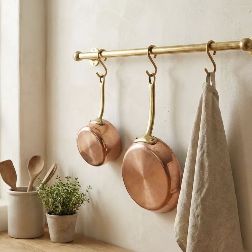 A beautiful and grounded kitchen scene showcasing two copper pans hanging from a classic horizontal brass rail. The rail is mounted above a smooth, light wood countertop that reflects a soft glow. To the side of the hanging pans, a simple ceramic pot holds an assortment of wooden mixing spoons, while another pot contains a thyme plant. A natural linen hand towel hanging gracefully beside them. Filled with warm, natural light, the entire image highlights a peaceful, minimalist, and non-toxic approach to kitchen design and mindful meal preparation.